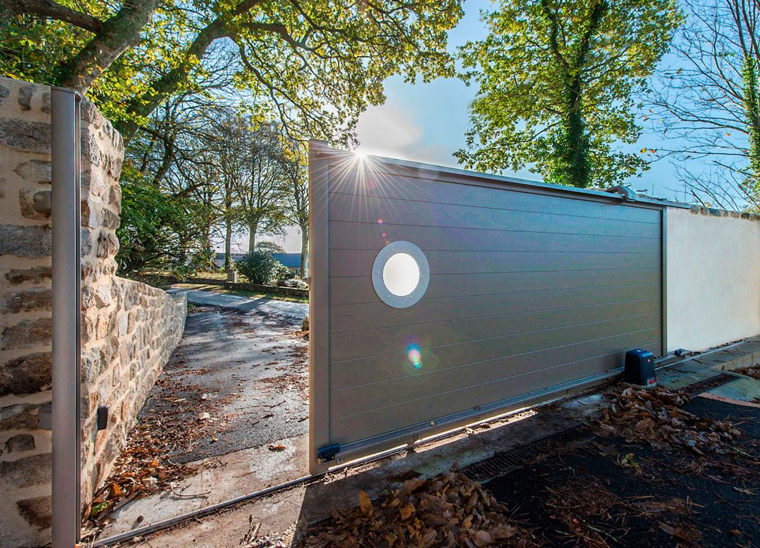 Portail coulissant gris-taupe à lames horizontales et hublot rond, mur pierre