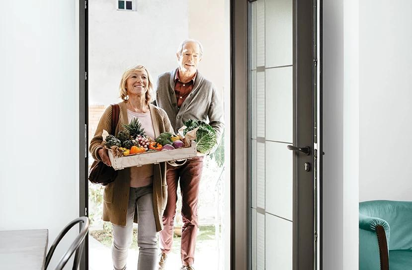 Porte d'entrée aluminium anthracite ouverte, couple portant une caisse de légumes