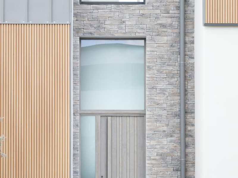 Porte d'entrée bois gris clair avec vitrage latéral et imposte, mur pierre