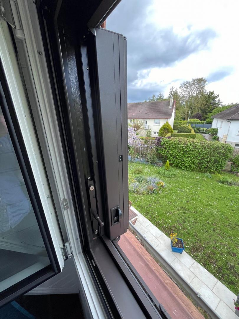 Volet pliant aluminium anthracite sur fenêtre ouverte, vue jardin et maison